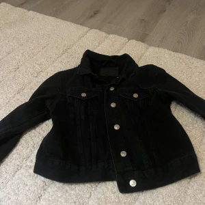 Svart jeansjacka med silvriga knappar - Snygg svart jeansjacka med klassisk krage och silvriga knappar framtill. Jackan har två bröstfickor med lock och knappar, samt långa ärmar. Perfekt för dig som vill ha en tidlös och cool look.