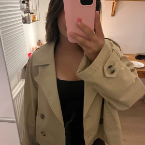 Beige croppad trenchcoat från H&M - Säljer en beige croppad trenchcoat från H&M med dubbelknäppning, stora slag och axelklaffar. Jackan har långa ärmar och snygga knappar vid ärmslut och axlar. Perfekt för lager-på-lager och ger en cool vibe till din outfit.