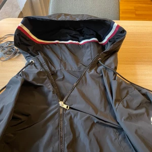 Svart vindjacka från Moncler - Säljer en svart vindjacka från Moncler med huva och snygga ränder i rött, vitt och blått på insidan av huvan. Jackan har dragkedja framtill och justerbar snörning vid huvan. Perfekt för blåsiga dagar och stilren look. Storlek 1/S Nypris ca 8000. Mitt pris 1649kr.