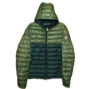 Moncler jacka - Snygg dunjacka från Moncler i två nyanser av grönt. Jackan har quiltad design, huva och dragkedja framtill. Klassisk Moncler-logga på ärmen och innerfoder med märkets etikett. Perfekt för dig som vill ha en lätt och stilren jacka med streetkänsla.