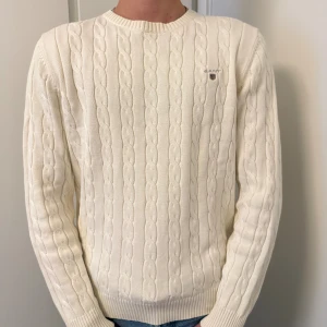Vit kabelstickad tröja från GANT - Säljer en klassisk vit kabelstickad tröja från GANT i storlek M. Tröjan har rund halsringning, ribbade muddar och diskret GANT-logga på bröstet. Perfekt för lager-på-lager och passar till jeans eller chinos.