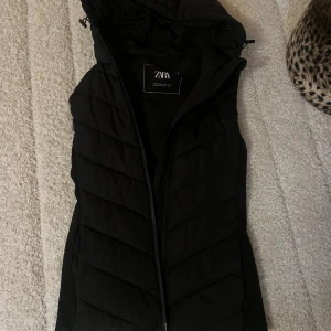Svart dunväst från Zara med huva - Snygg svart dunväst från Zara med huva och dragkedja framtill. Västen har en quiltad design och är perfekt för lager-på-lager-stil. Passar dig som gillar en sportig och trendig look.
