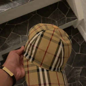 Rutig keps från Burberry - Snygg keps från Burberry i klassiskt beige rutigt mönster med inslag av svart, vitt och rött. Kepsen har böjd skärm och justerbar rem baktill. Perfekt accessoar för att lyfta din outfit med en lyxig touch.
