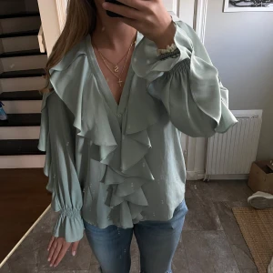 Ljusgrön blus med volanger - Supersöt ljusgrön blus med stora volanger framtill och längs ärmarna. Blusen har v-ringning, långa ärmar med resår vid handlederna och en flowy passform. Perfekt för dig som gillar romantisk och trendig stil.
