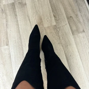 Svarta mocka cowboy boots med klack - Snygga svarta cowboy boots i mocka med spetsig tå och markerad klack. Stövlarna har dragkedja på sidan och en cool western-inspirerad form upptill. Perfekta för att lyfta vilken outfit som helst.