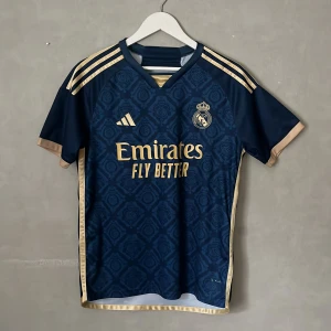 Real Madrid tröja med en grafisk design - Snygg Real Madrid fotbollströja med Vini Jr och nummer 7 på ryggen. Marinblå med guldiga detaljer och mönstrad framsida, Adidas-logga och klubbmärke. Kortärmad med guldfärgade ränder på axlarna och kanter. Perfekt för dig som älskar fotboll och stil.