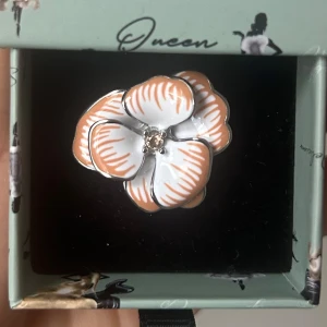 Ioaku Santorini Fleur - Superfin ring i form av en blomma med vita och ljusorange kronblad och en liten glittrande kristall i mitten. Ringen har en cool och unik design som verkligen sticker ut och passar perfekt om du vill ha något extra på handen. Från Ioaku i färgen Cappuccino, storlek 7. Ordinarie pris 699 kr