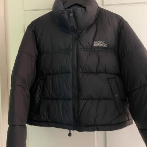 Svart pufferjacka från Pacific Republic - Säljer en svart pufferjacka från Pull&Bear. Snygg lite kortare streetwear jacka. Vet ej fraktkostnaden.