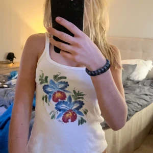 Vit linne med blommor❤️ - Vit linnetopp från On the Beach som jag köpt i spanien,  med smala axelband och tryck av stora blå och röda blommor framtill. Perfekt för varma dagar och ger en fräsch, somrig vibe. Mjuk bomullskänsla och enkel att matcha med shorts eller kjol. Storlek L med passar mig som är xs