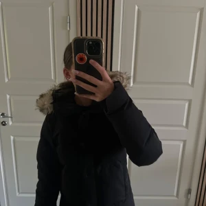 Svart pufferjacka - Varm och snygg svart vadderade vinterjacka med avtagbar huva och fuskpälsdetalj. Jackan stängs med dragkedja och tryckknappar framtill, har två sidflickor samt en praktisk innerficka för extra förvaring. Perfekt för kalla dagar- både strilen och funktionell.