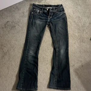 Premiere jeans med massa detaljer på fickan - Mörkblå jeans från Premiere med snyggt slitna partier och kontrastsömmar. Baksidan har broderade fickor och dekorativa detaljer med fransar och nitar. Klassisk femficksmodell med låg midja och bootcut-ben. En nit har tappat på fickan där bak!
