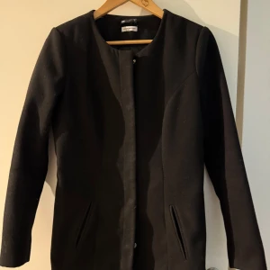 Svart kappa från JDY - Stilren svart kappa från jacqueline de yong med rak passform och diskret knäppning framtill. Jackan har två fickor och är helsvart utan krage, vilket ger en clean look. Perfekt för dig som gillar minimalistisk stil. En knapp har lossat.