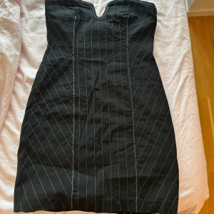 Svart axelbandslös randig kortklänning - Snygg svart axelbandslös kortklänning med vita ränder. Klänningen har en v-ringad överdel och är figurnära med markerade sömmar.