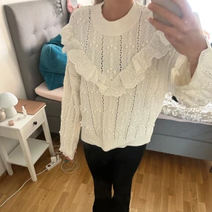 Vit stickad tröja med volang från H&M - Superfin vit stickad tröja från H&M med broderade volanger och hålmönster. Tröjan har lång ärm, rund halsringning och detaljer med små bollar och spetsvolang både fram och bak. Perfekt för dig som gillar romantisk och söt stil!