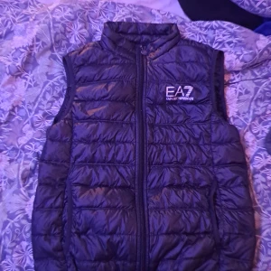 Svart dunväst från EA7 Emporio Armani - Snygg svart dunväst från EA7 Emporio Armani med quiltad design och hög krage. Västen har en tydlig logga på bröstet och dragkedja framtill med EA7-detalj. Perfekt att slänga över en hoodie för en cool look.