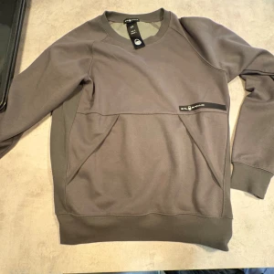 Grå sweatshirt från Sail Racing - Säljer en stilren grå sweatshirt från Sail Racing med diskret logga på bröstet. Tröjan har rund hals, långa ärmar och praktiska fickor framtill. Tillverkad i mjuk bomullsmix som är skön att bära. Perfekt för en chill och sportig look.