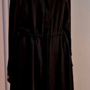 Svart långärmad skjortklänning från H&M - Snygg svart skjortklänning från H&M i storlek 40. Modellen har lång ärm, v-ringad hals och ett markerat midjeparti med snörning för en schysst siluett. Perfekt för dig som gillar stilrena och enkla plagg med lite edge.Tyvärr är klänningen för stor för dockan som har stl 36.Annars sitter den perfekt.Oanvänd p.g.a felköp
