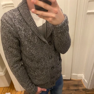 Grå stickad cardigan från Cedar Wood State - Säljer en snygg grå stickad kofta från märket Cedar Wood State. Koftan har en klassisk design med knappar framtill och långa ärmar. Perfekt för kyligare dagar och ger en stilren look. Passar bra till jeans eller chinos.