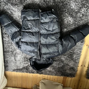 Svart dunjacka med huva - Säljer en svart dunjacka med huva och dragkedja. Jackan har en pufferdesign som ger en skön och varm känsla. Perfekt för kyliga dagar.