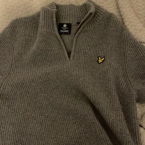 Grå stickad tröja från Lyle & Scott - Säljer en stilren grå stickad tröja från Lyle & Scott med dragkedja vid halsen. Tröjan har en broderad logotyp på bröstet och är perfekt för kyligare dagar. Passar både till vardags och mer uppklädda tillfällen.
