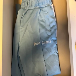 Ljusblå byxor från Palm Angels - Snygga ljusblå byxor från Palm Angels med elastisk midja och vita sidopaneler. Perfekta för en avslappnad stil. Byxorna har en broderad logga på benet och praktiska fickor.