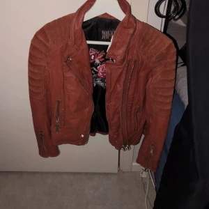 Röströd skinnjacka med dragkedjor - Snygg brun skinnjacka med coola dragkedjedetaljer och quiltade axlar. Jackan har en klassisk bikerstil med flera fickor och en asymmetrisk dragkedja framtill. Perfekt för en edgy look!