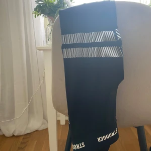 Svarta leggings från Stronger - Snygga svarta leggings från Stronger med vita prickiga ränder. Perfekta för träning med en tight passform och elastiskt material som ger bra rörelsefrihet. Texten 'STRONGER' finns tryckt vid midjan.