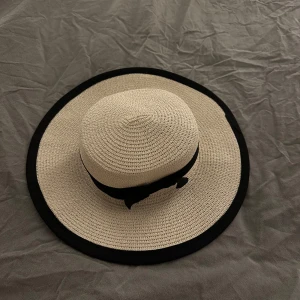 Beige solhatt med svart kant - Elegant/classy beige solhatt med bred svart kant och en svart rosett. Perfekt för soliga dagar. Tillverkad i ett flätat material. Endast testad 