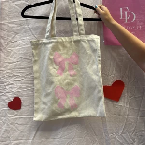 Beige shoppingväska med rosa rosetter - Vi är ett UF företag som säljer tygväskor med handmålade tryck på framsidan. Perfekt för att bära med sig det nödvändigaste med stil.