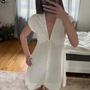 Vit kortklänning med knappar - Säljer en söt vit kortklänning med knappar framtill. Klänningen har en v-ringad design och korta ärmar, perfekt för varma dagar.🤍Ej använd, endast provad. Nypris 650kr