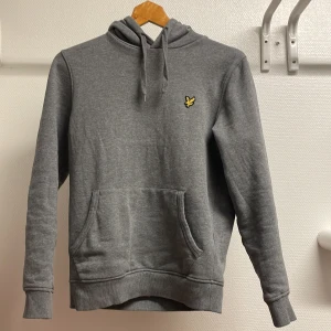 Grå hoodie från Lyle & Scott - Säljer en stilren grå hoodie från Lyle & Scott med deras ikoniska gula logga på bröstet. Tröjan har en klassisk känguruficka och justerbar huva med dragsko. Perfekt för en avslappnad look.