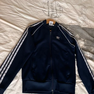 Mörkblå träningsjacka från Adidas - Säljer en snygg mörkblå träningsjacka från Adidas med de klassiska tre vita ränderna längs ärmarna. Jackan har dragkedja framtill och en liten Adidas-logga på bröstet. Perfekt för sportiga aktiviteter eller en avslappnad stil.