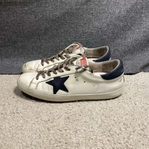 Vita sneakers med stjärndetalj från Golden Goose - Snygga vita sneakers från Golden Goose med en distinkt mörkblå stjärna på sidan. Skorna har en klassisk snörning och en mörkblå häl. Dom har däremot gott upp lite i hälen. Priset kan gå ner 