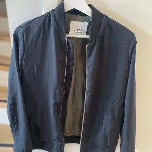 Svart bomberjacka från Stay - Snygg svart bomberjacka från Stay med dragkedja framtill. Jackan har en klassisk design med ribbade muddar vid ärmslut och nederkant.