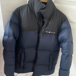 Mörkblå dunjacka från Sail Racing - Säljer en snygg mörkblå dunjacka från Sail Racing. Jackan har en hög krage och dragkedja framtill. Den är perfekt för kyliga dagar och har en stilren design med logotyp på bröstet och ryggen. Mittersta sömmen på ryggen har lossat!