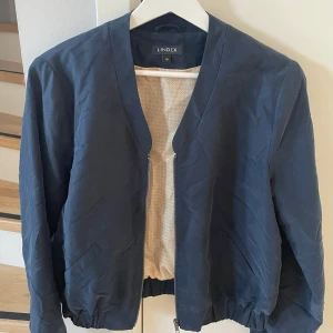 Mörkblå bomberjacka från Lindex - Mörkblå bomberjacka från Lindex med dragkedja framtill. Jackan har en klassisk design med ribbade muddar vid ärmslut och nederkant. 