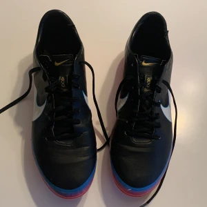 Nike Mercurial fotbollsskor - Snygga Nike Mercurial fotbollsskor i svart med blå och rosa detaljer. Skorna har snörning och en stilren design med en vit Nike-logga på sidan. Perfekta för fotbollsplanen med en greppvänlig sula.