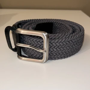 Grått flätat bälte med silverfärgat spänne - Snyggt grått flätat bälte med ett stilrent silverfärgat spänne. Bältet har svarta läderdetaljer vid spännet som ger en extra touch av elegans. Perfekt för att ge din outfit en modern och trendig look.