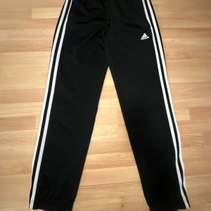 Svarta träningsbyxor från Adidas - Säljer ett par klassiska svarta träningsbyxor från Adidas med vita ränder längs sidorna. Byxorna har en bekväm passform och är perfekta för sport och fritid. Adidas-loggan är tryckt på ena benet.