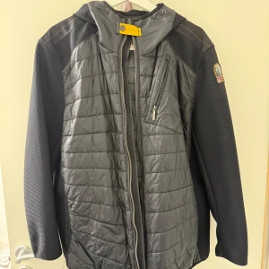 Svart jacka från Parajumpers - Säljer en svart jacka från Parajumpers med quiltad design och huva. Jackan har en dragkedja framtill och en bröstficka med dragkedja. Den har ett märke på ärmen och orangea detaljer invändigt. Perfekt för kyligare dagar.