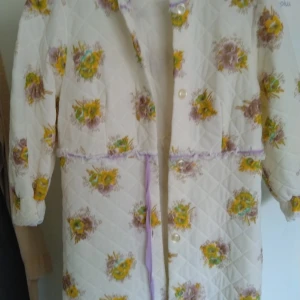 Blommig quiltad kappa från David Flow morgon Rock er - Charmig quiltad kappa från David Flower med blommigt mönster i gult och lila. Kappan har långa ärmar och knappar framtill. Perfekt för en unik och färgglad stil.