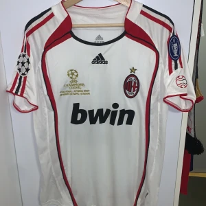 Vit AC Milan fotbollströja från Adidas - Säljer en vit AC Milan fotbollströja från Adidas med röda och svarta detaljer. Tröjan har korta ärmar och klubbens emblem på bröstet. Den har även trycket 'KAKA' och nummer 22 på ryggen. Perfekt för fans av laget!