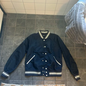 Marinblå bomberjacka från GANT - Säljer en snygg marinblå bomberjacka från GANT med vita detaljer. Jackan har långa ärmar och stängs med knappar framtill. Den har ribbade muddar och krage med vita ränder samt GANT-logga på ryggen. Perfekt för en stilren look!