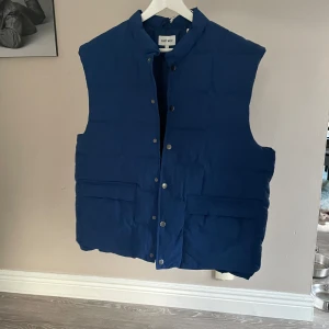 Blå dunväst från East West - Snygg blå dunväst från East West med knappar framtill. Perfekt för kyligare dagar och har en stilren design med praktiska fickor.