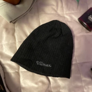 Svart mössa från Ullmax - Snygg och enkel svart mössa från Ullmax. Perfekt för kyliga dagar med sin stilrena design och bekväma passform. Broderad logga framtill ger en extra touch.  