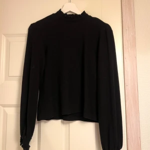 Svart långärmad topp med hög krage - Elegant svart topp med hög krage och långa ärmar. Perfekt för en stilren look. Toppen har en knappdetalj i nacken för enkel på- och avtagning.