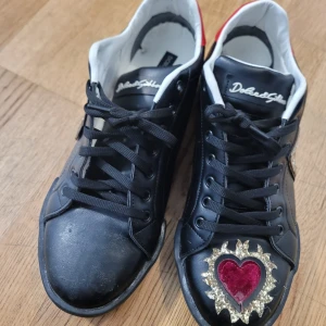 Svarta sneakers från Dolce & Gabbana - Snygga svarta sneakers från Dolce & Gabbana med glittrande detaljer. Skorna har ett hjärta i rött och guld på tån och stora DG-bokstäver i guldglitter på sidan. Tillverkade i Italien med snörning framtill. Jag använde skorna bara en gångoch dom är i väldig bra skicka. Jag kan frakta samma dag. Det finns en del som jag tog bort men kan limmas på  enkelt.