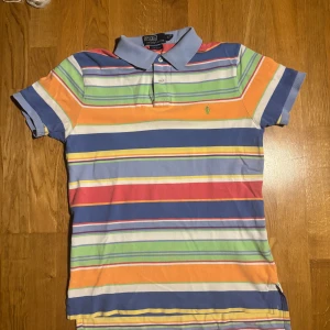 Flerfärgad randig pikétröja från Ralph Lauren - Säljer en färgglad pikétröja från Ralph Lauren med ränder i blått, grönt, orange, gult och vitt. Tröjan har en klassisk krage och korta ärmar. Perfekt för en avslappnad stil med en touch av färg.