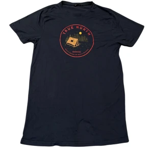Svart t-shirt från True North - Säljer en svart t-shirt från True North med ett coolt tryck av ett tält och texten 'True North' i rött på bröstet. Perfekt för en avslappnad stil. T-shirten är kortärmad och gjord i mjukt material.