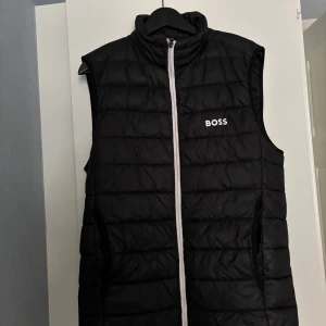 Svart dunväst från Hugo Boss - Snygg svart dunväst från Boss med dragkedja framtill. Västen har en stilren design med märkets logga på bröstet. Perfekt för lager-på-lager under kyligare dagar. Nypris 1800 säljs för 550 kr. 